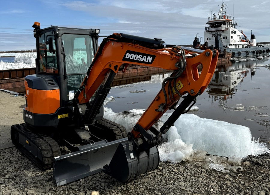 Мини-экскаватор Doosan припаркован возле частично замерзшей реки с лодками на заднем плане. Промтех - механизация