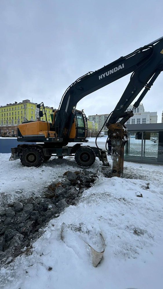Экскаватор бурит заснеженный грунт на строительной площадке зимой. Промтех - механизация